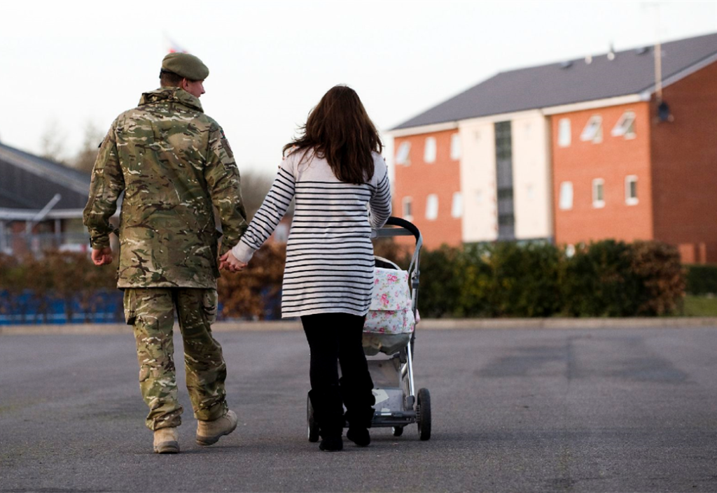 Soldier in fatigues and female partner hold hands as they walk towards some low-rise flats. The mum is pushing a pram with her free (right) hand